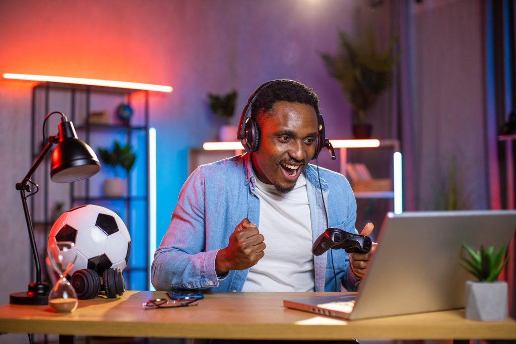 Man wearing a headset and denim shirt celebrates while gaming on a laptop, holding a game controller.