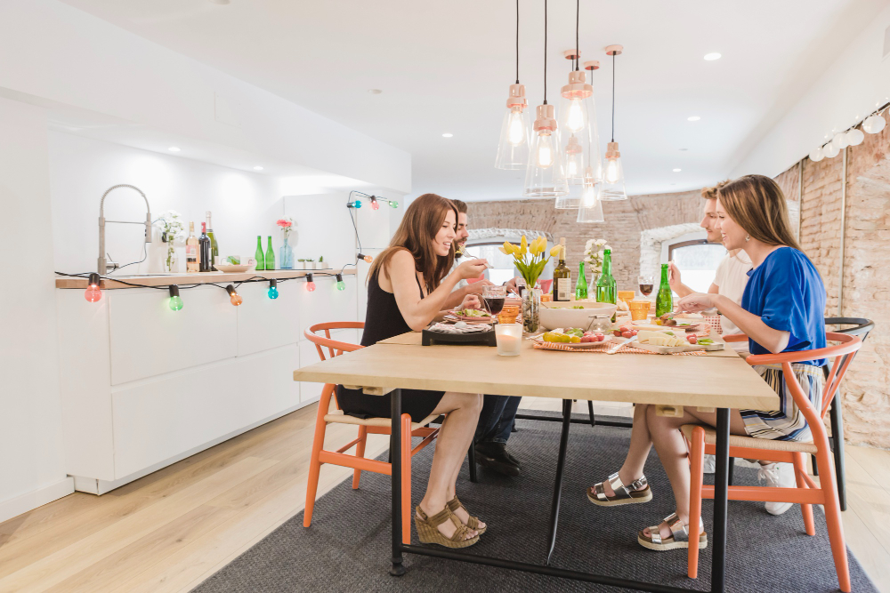 Group of four friends dining at a wooden table in a bright, open kitchen, sharing plates and drinks from green bottles.