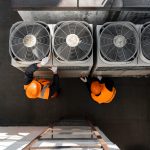 Overhead view of two maintenance workers in orange safety vests and hard hats servicing three large outdoor HVAC units on a rooftop.
