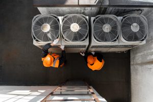 Overhead view of two maintenance workers in orange safety vests and hard hats servicing three large outdoor HVAC units on a rooftop.