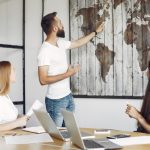 Bearded man presenting a world map on a wooden panel wall to two colleagues in a meeting room.