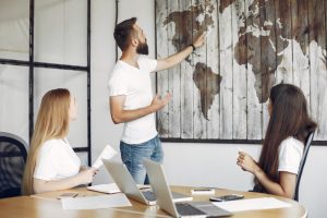 Bearded man presenting a world map on a wooden panel wall to two colleagues in a meeting room.