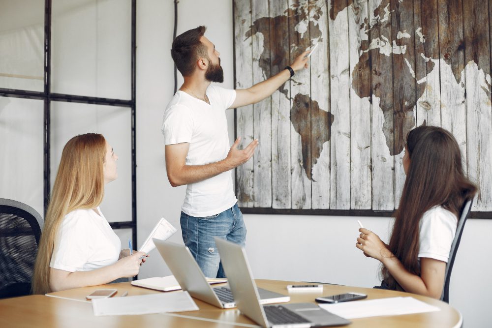 Bearded man presenting a world map on a wooden panel wall to two colleagues in a meeting room.