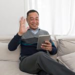 Man sitting on a light couch at home, smiling and waving while holding a tablet.
