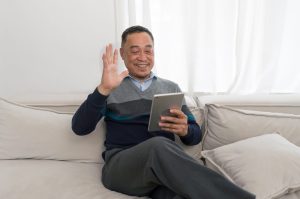 Man sitting on a light couch at home, smiling and waving while holding a tablet.