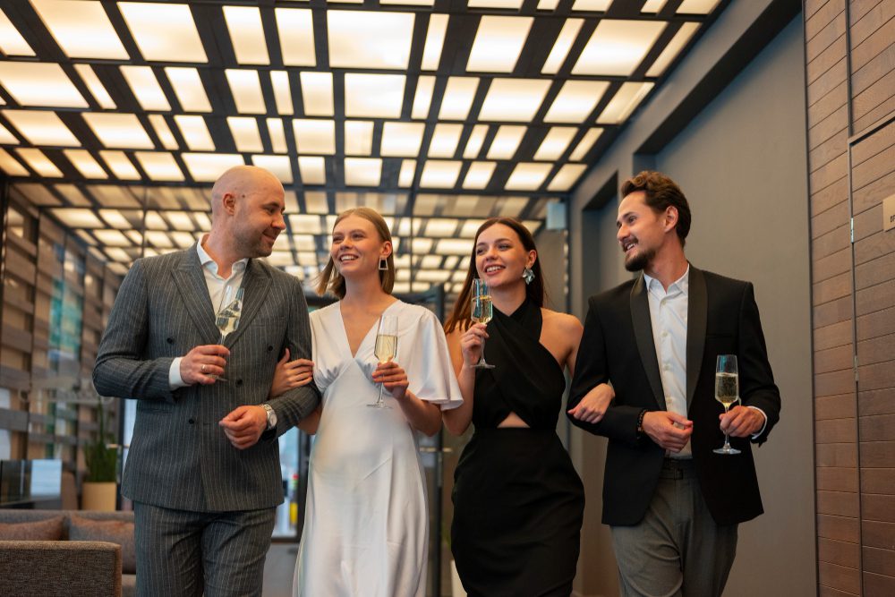 Group of four friends in formal attire walking down a contemporary hallway, each holding a champagne glass and smiling.