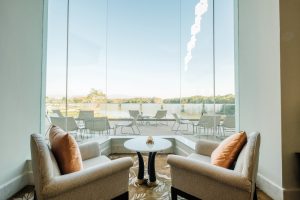 Two beige armchairs with orange cushions flank a round white table in a glass-walled lounge overlooking a patio and lake.
