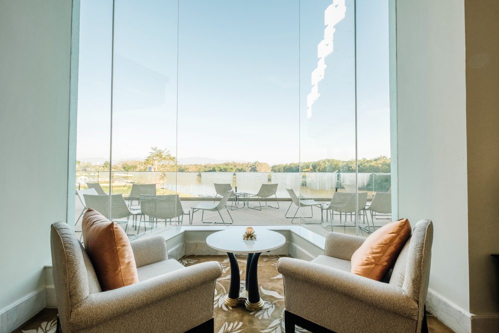 Two beige armchairs with orange cushions flank a round white table in a glass-walled lounge overlooking a patio and lake.