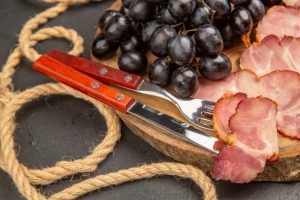 Charcuterie scene: red-handled knife and fork on a wooden board with grapes and sliced ham.