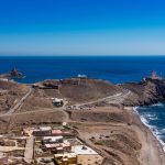 Coastal hillside town with a winding road toward a rocky headland and a lighthouse overlooking the blue sea.