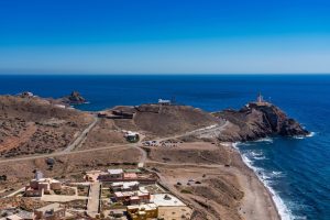 Coastal hillside town with a winding road toward a rocky headland and a lighthouse overlooking the blue sea.