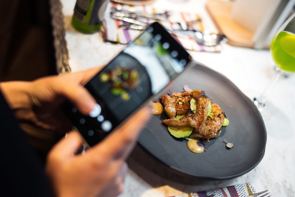Person holds a smartphone to photograph a plated dish on a dark triangular plate with green accents and sauce dollops.