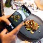 Person holds a smartphone to photograph a plated dish on a dark triangular plate with green accents and sauce dollops.
