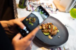 Person holds a smartphone to photograph a plated dish on a dark triangular plate with green accents and sauce dollops.