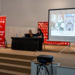 Two presenters sit at a long black-draped table on a stage, presenting with a projector screen showing a collage and the words 'TORRIJOS: UNA PASION QUE SE TRANSFORMA EN ARTE'. Beside them, a red Bienal Viral banner stands on stage.