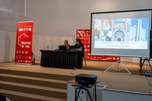 Two presenters sit at a long black-draped table on a stage, presenting with a projector screen showing a collage and the words 'TORRIJOS: UNA PASION QUE SE TRANSFORMA EN ARTE'. Beside them, a red Bienal Viral banner stands on stage.