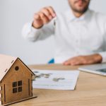 Man in a white shirt hands over a key in front of a wooden house model on a desk with papers and a laptop nearby, symbolizing a real estate transaction.