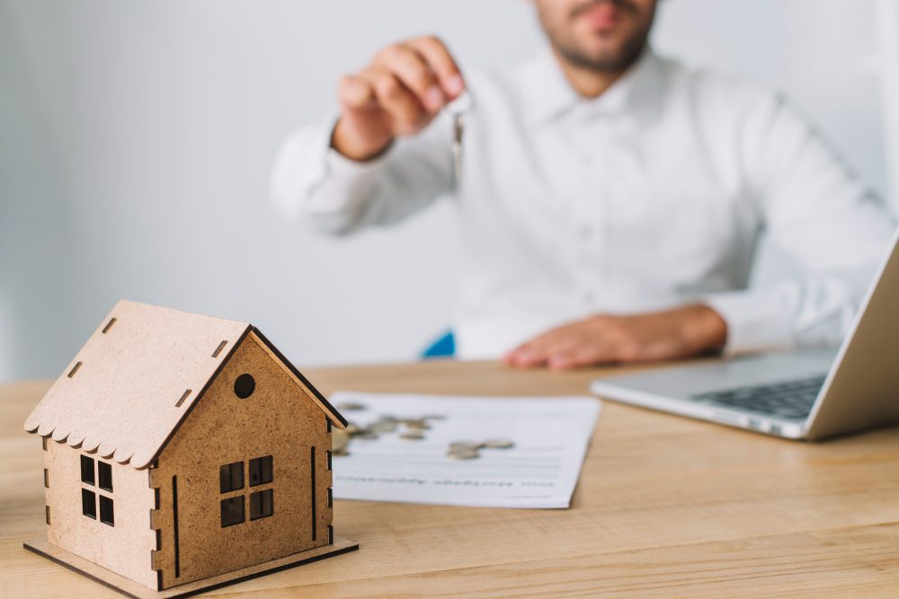 785139 - Soms Granados | Diario Digital XXI Man in a white shirt hands over a key in front of a wooden house model on a desk with papers and a laptop nearby, symbolizing a real estate transaction.